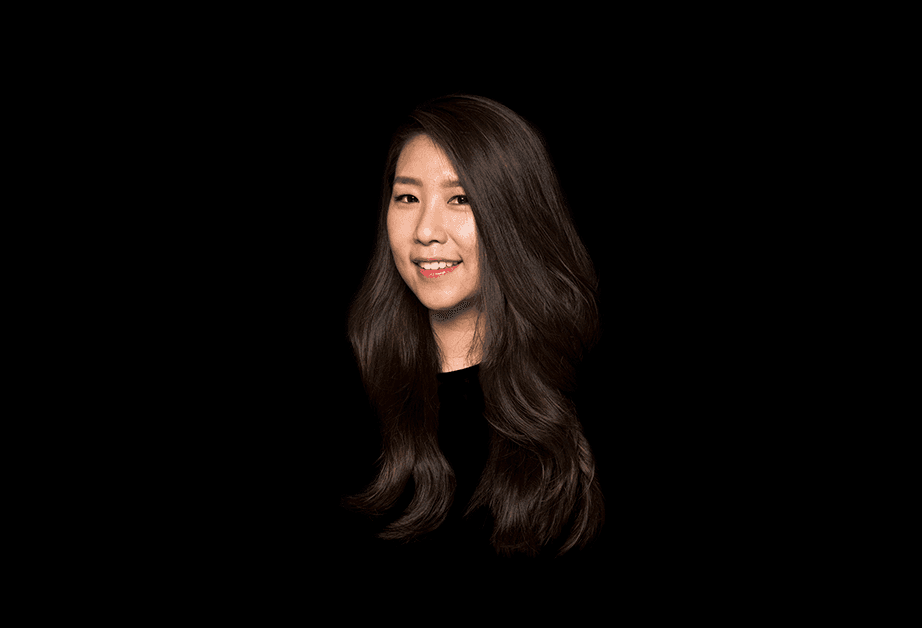 A woman with long, dark brown hair is shown against a solid black background. She is looking directly at the camera and smiling slightly. Her hair falls over her shoulders and partially obscures the black top she is wearing. The lighting is focused on her face and upper body, creating a clear contrast with the dark background. No other objects or text are visible in the image.