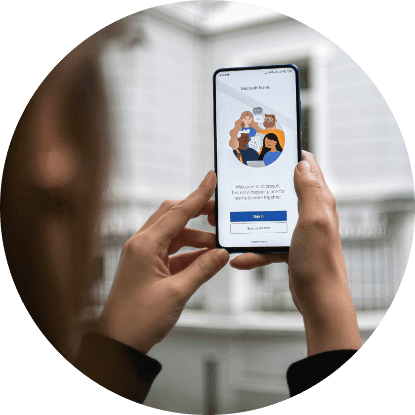 A person holding a smartphone displaying the Microsoft Teams welcome screen, featuring illustrations of diverse people collaborating. The background shows a white building.