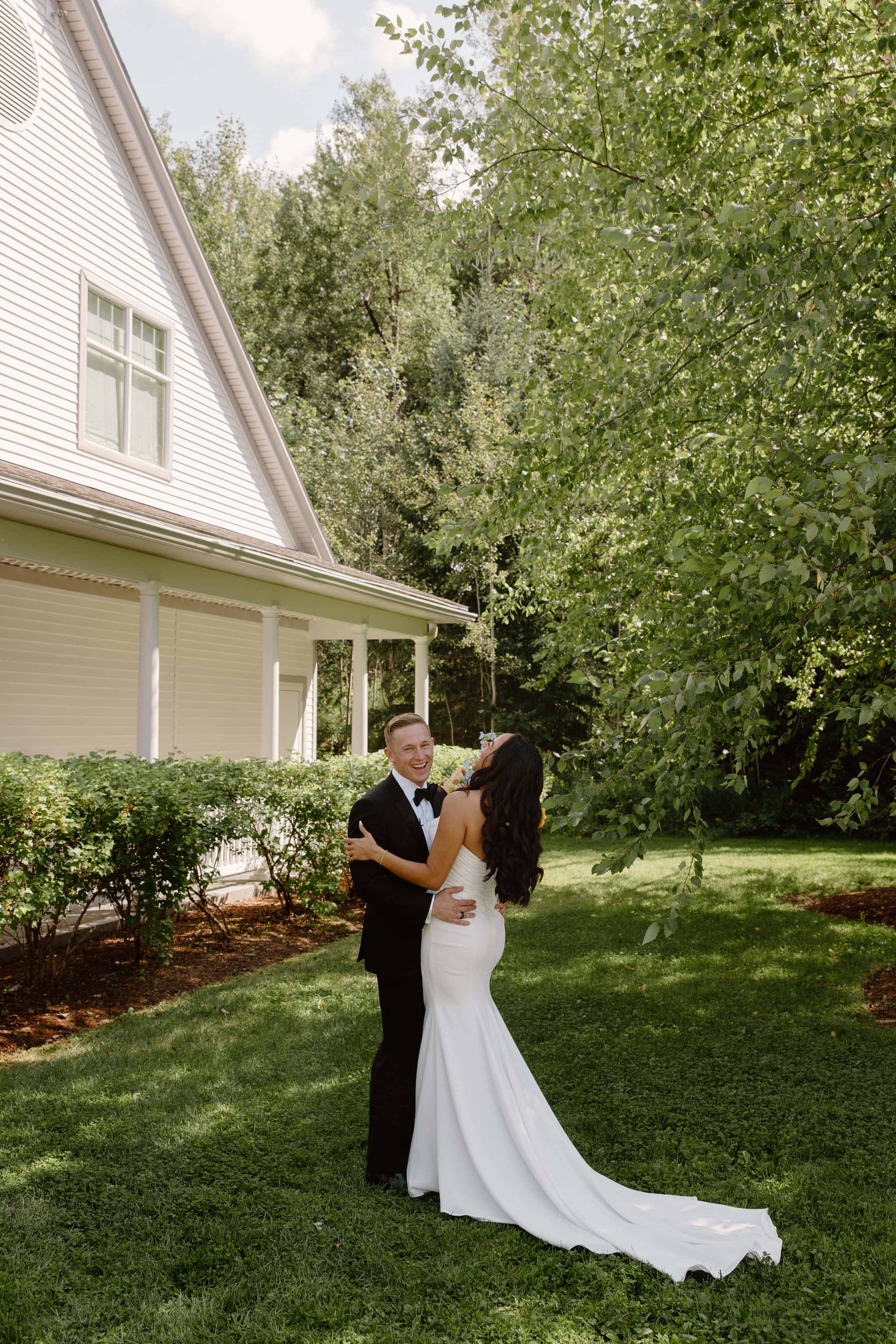  A photo of a couple standing in a grassy area outside a white house. The man wears a tuxedo and the woman wears a white dress.