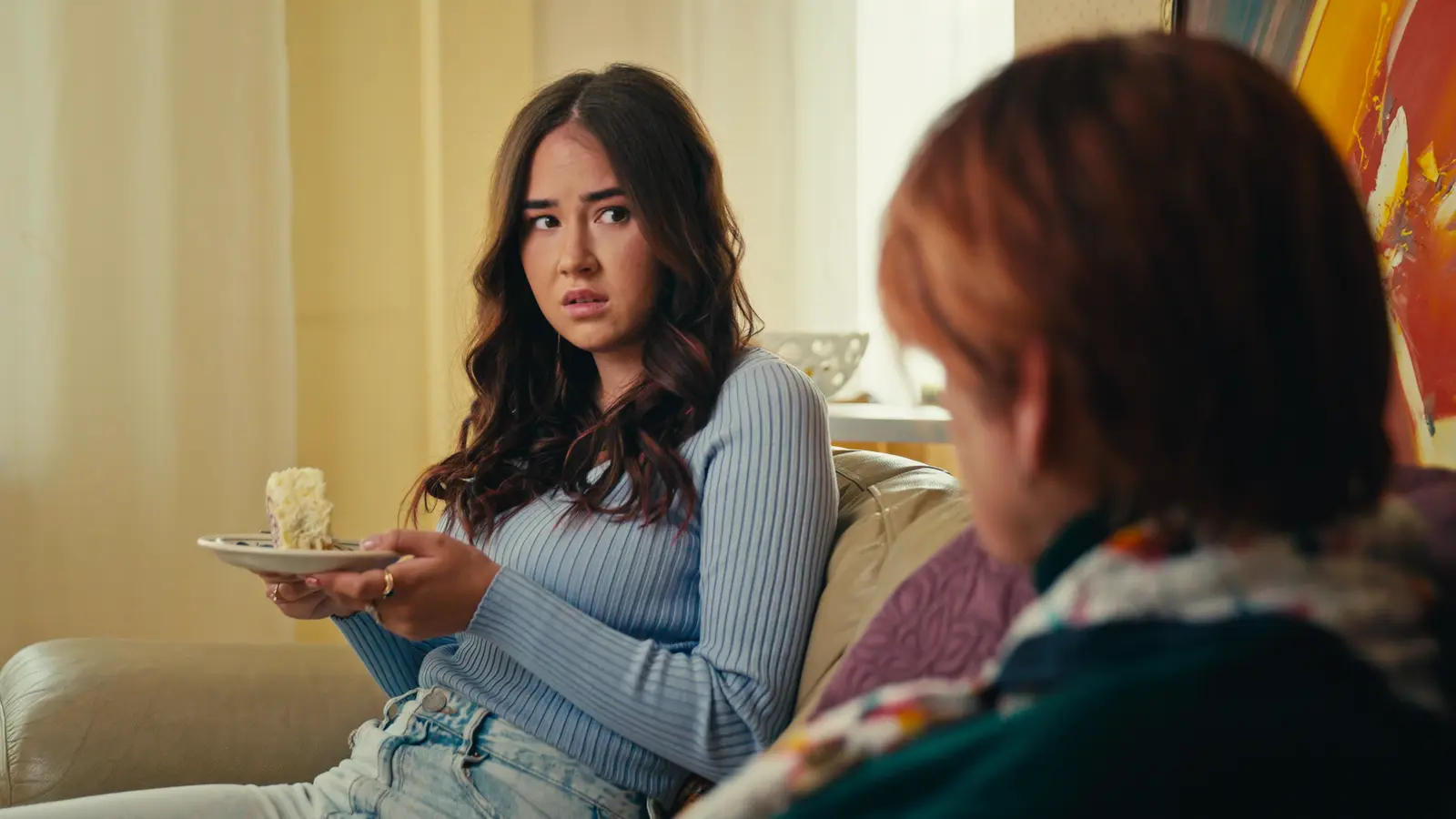 Woman holding a plate of cake looking uneasy beside an older woman on a couch