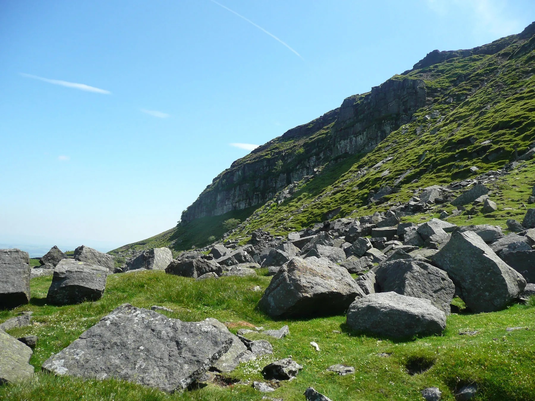 Rocky mountain hillside with scattered large boulders on green grass slope under clear blue sky, scenic hiking terrain