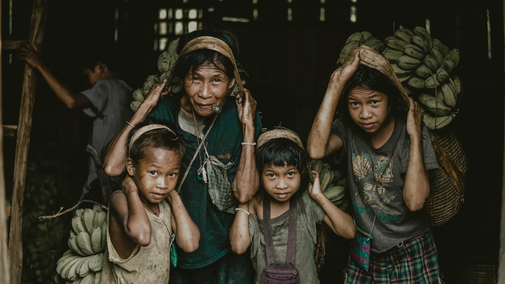 A family of four, including an elderly woman and three children, carrying large baskets of bananas on their backs, likely indicative of their work in agriculture or fruit harvesting. The image captures the resilience and labor of rural life.