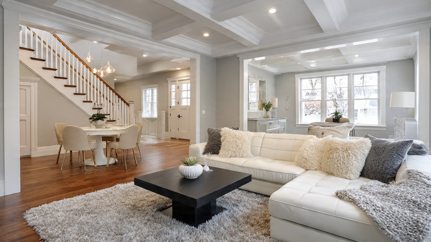 Modern living room with white sofa, fluffy cushions, and a black coffee table on a gray rug. Nearby are stairs, a dining area, and a bright kitchen.