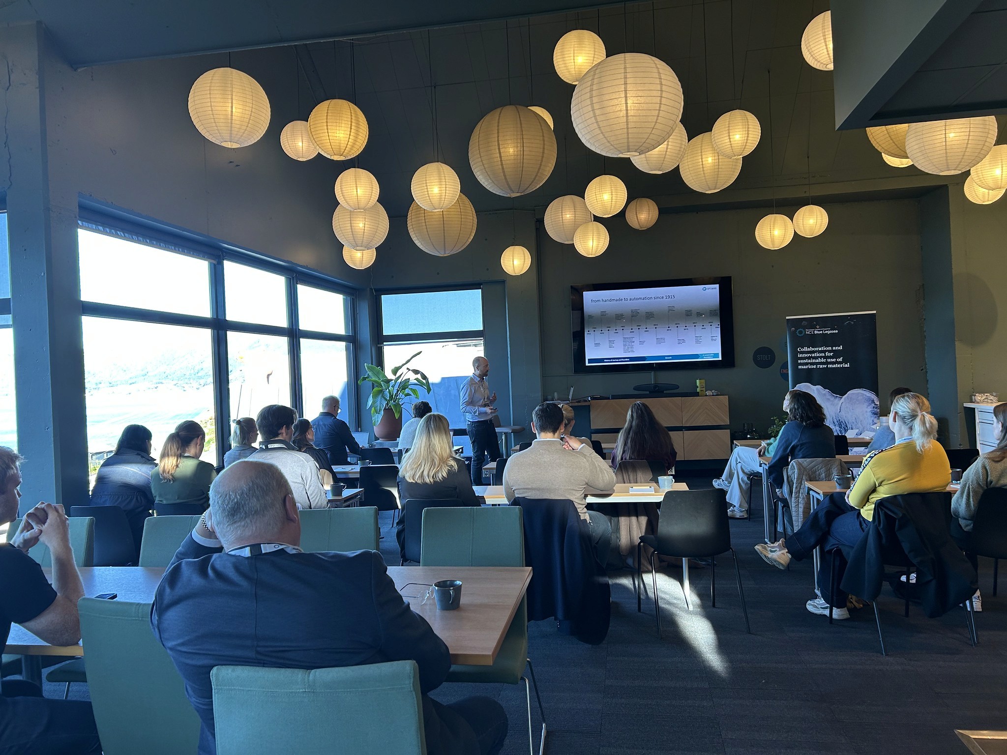 A spacious room filled with people sitting at tables, with large windows and pendant lights overhead.