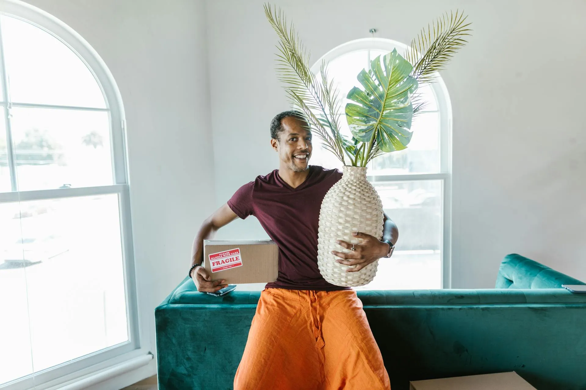Smiling man holding a large decorative plant vase and a fragile moving box while sitting on a green sofa.