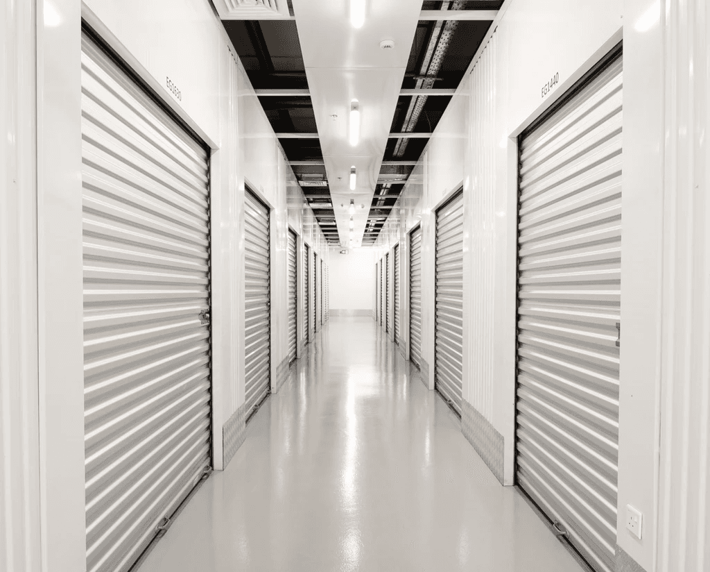 A brightly lit hallway with rows of closed, metallic storage unit doors on both sides. The polished concrete floor reflects the overhead lights, creating a sterile and orderly atmosphere.