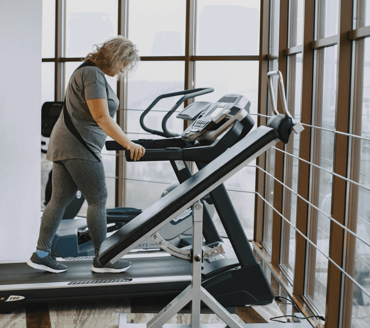 Woman in grey workout gear walks on a treadmill in a bright gym with floor-to-ceiling windows.