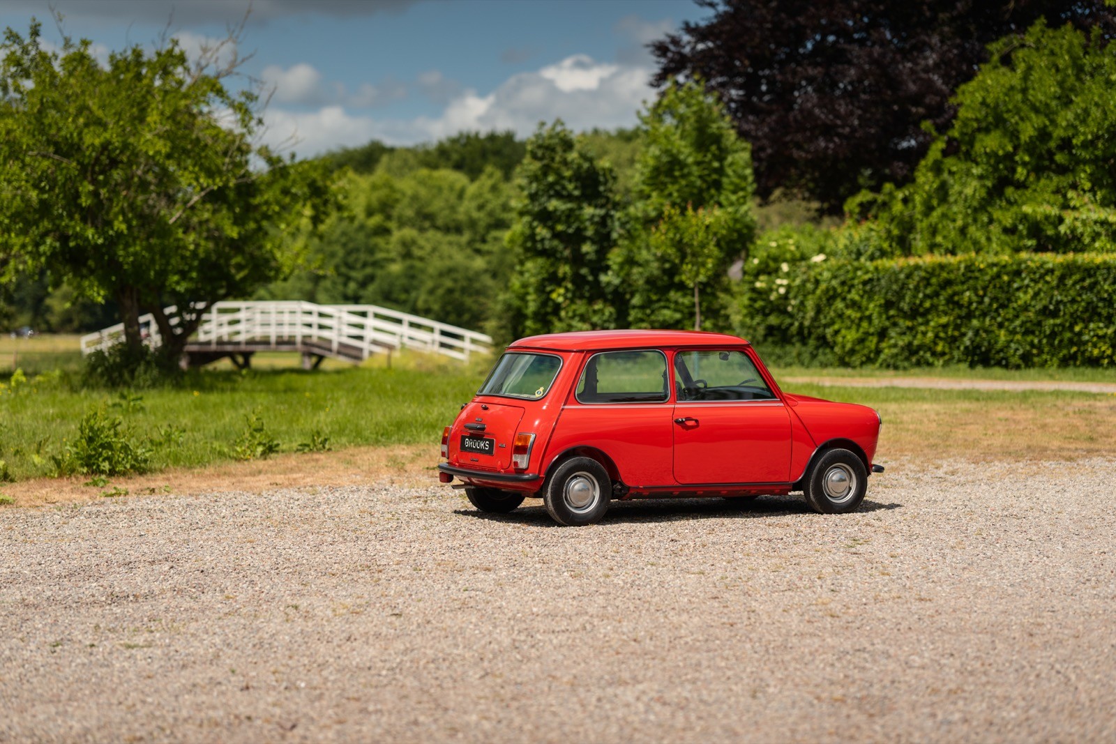 A vintage red car parked on a sunny day in a green, natural setting with trees in the background.