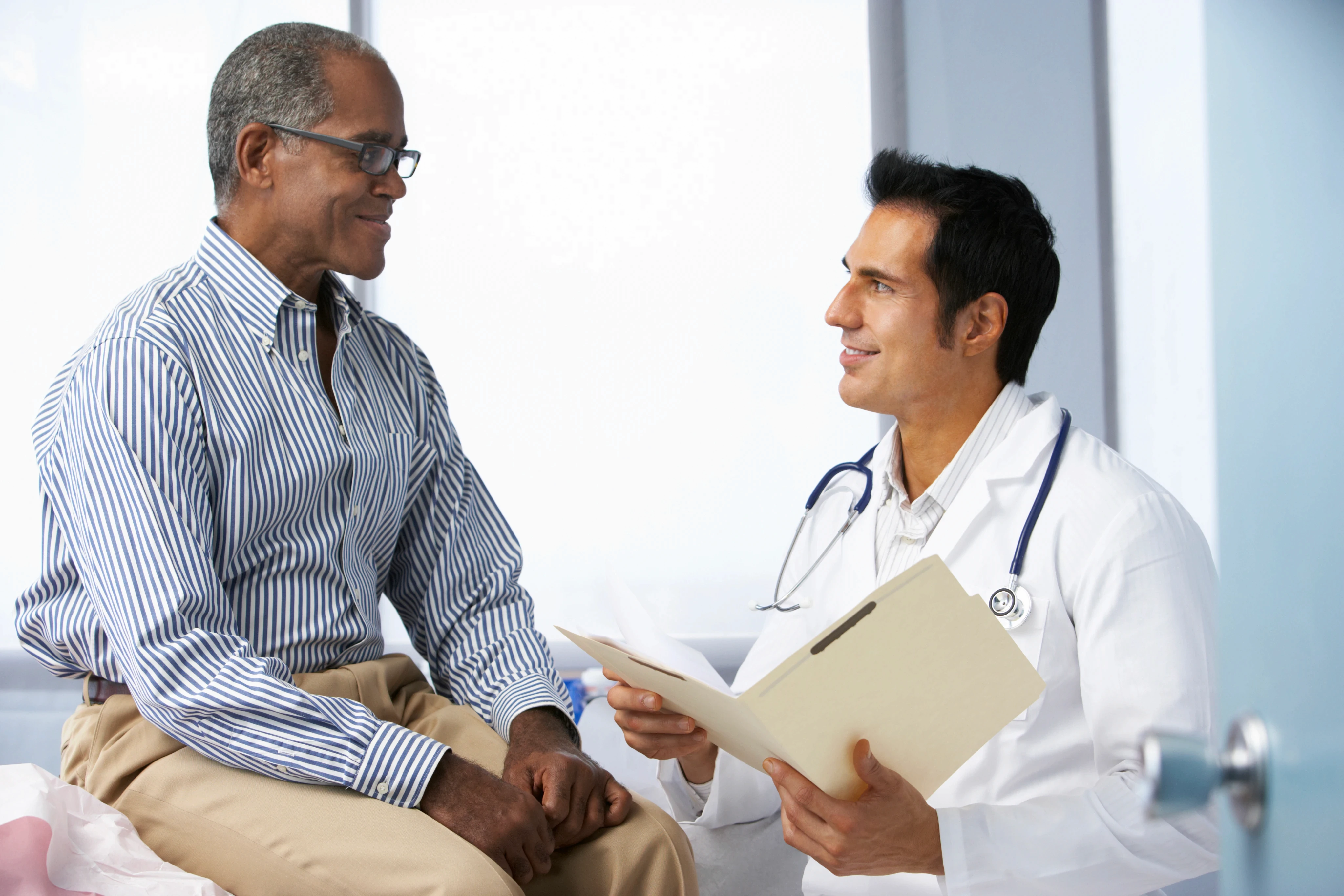 A doctor in a white coat discusses with an elderly man sitting on an exam table. Both are smiling, conveying a sense of trust and comfort in a bright clinic.