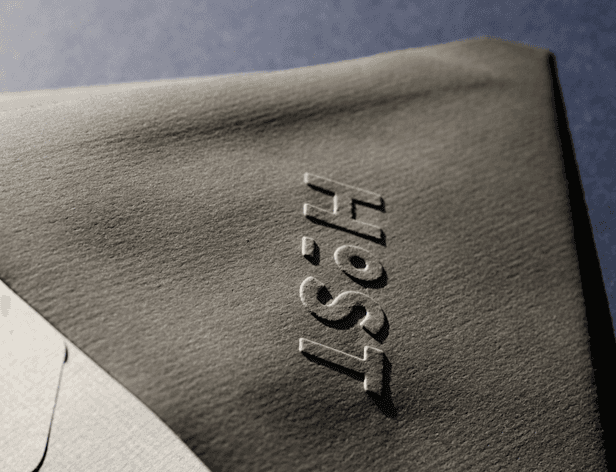 A close-up view of a dark gray fabric surface with the word "HOST" embossed in lighter gray capital letters. The fabric has a slightly textured appearance and a subtle shadow is cast to its right. A portion of a lighter-colored fabric is visible in the bottom left corner. The background is a darker gray tone.