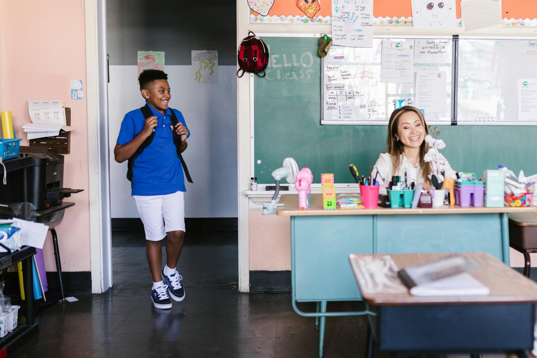 A smiling educator kneeling down to have a supportive one-on-one conversation with a young student at their desk.