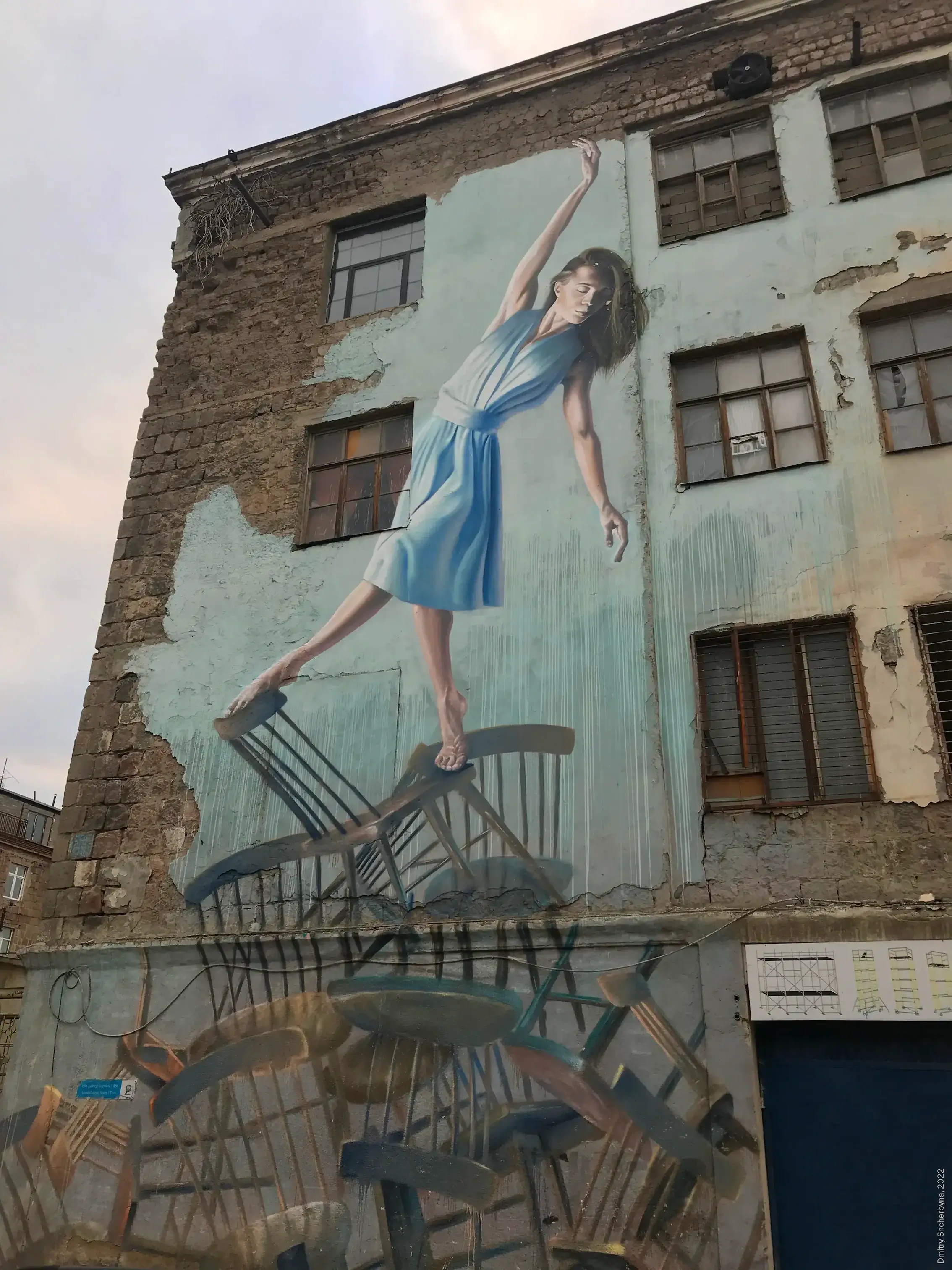 Photorealistic mural of a young woman in a light blue dress balancing gracefully on a toppling pile of wooden chairs and tables, painted on a weathered brick building facade.