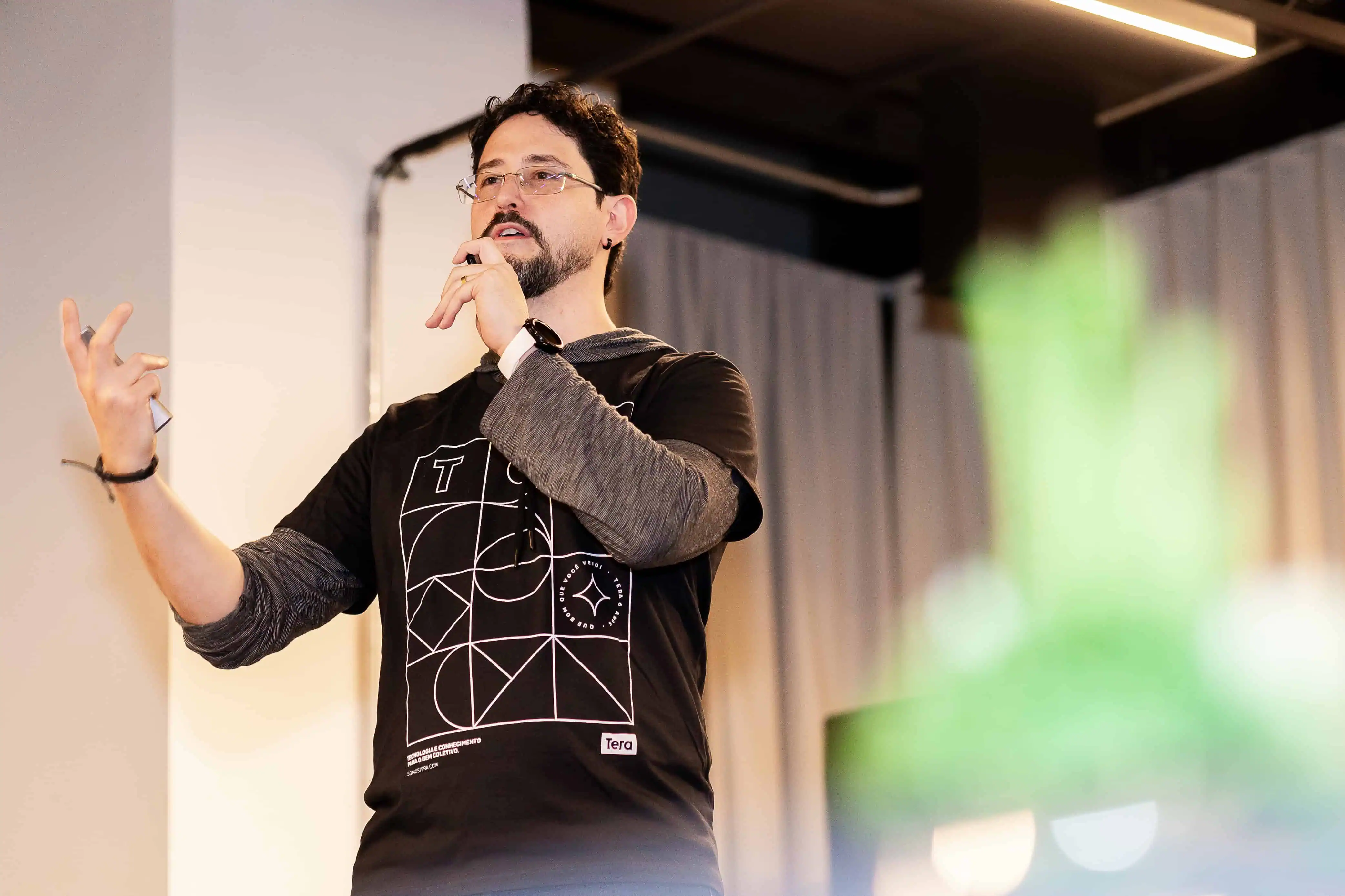 Uma pessoa está em pé no palco falando ao microfone, vestindo uma camiseta preta, com uma planta em primeiro plano.
