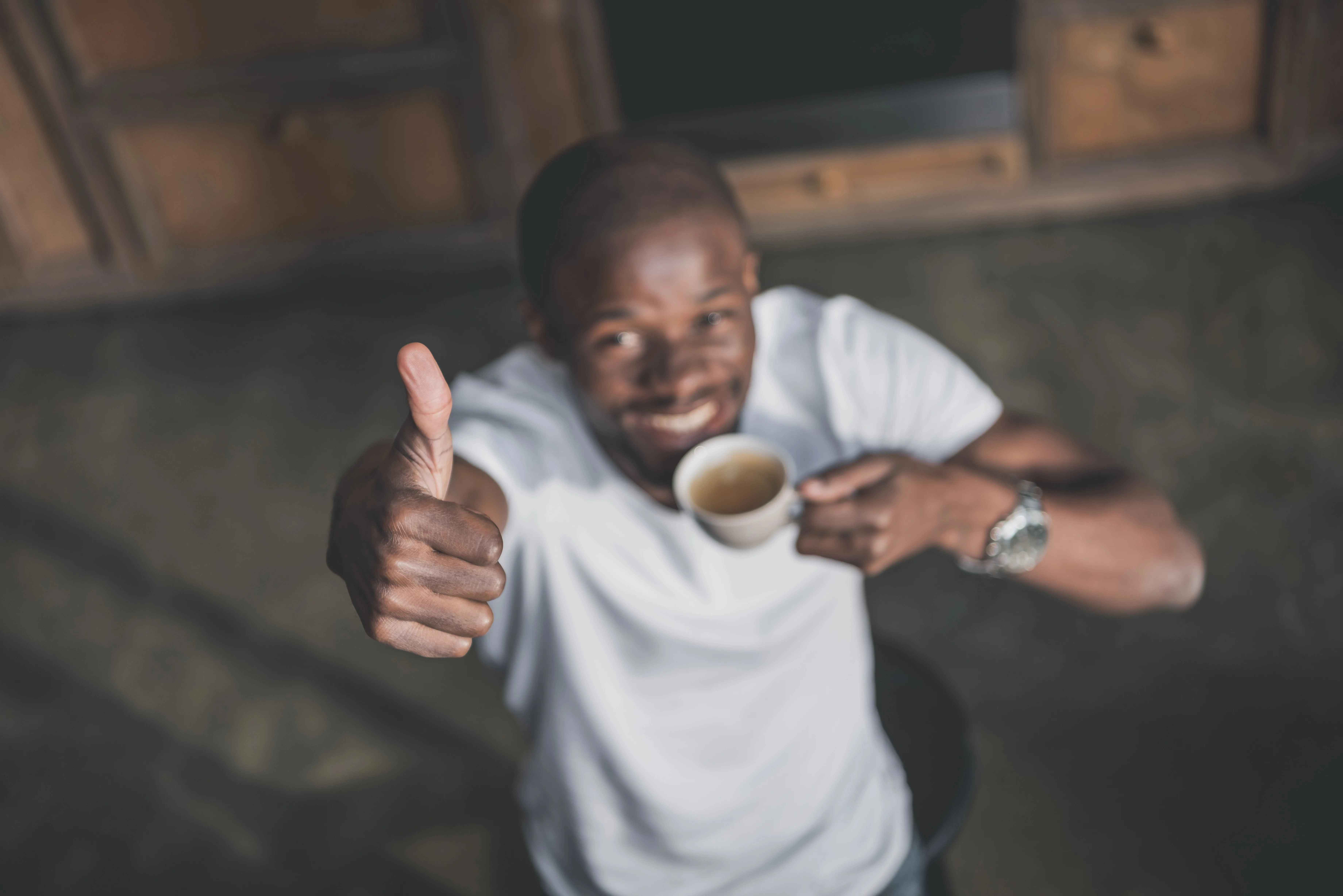 A elevated photo of a ethnic man holding a cup of coffee, smiling and giving a thumbs up.
