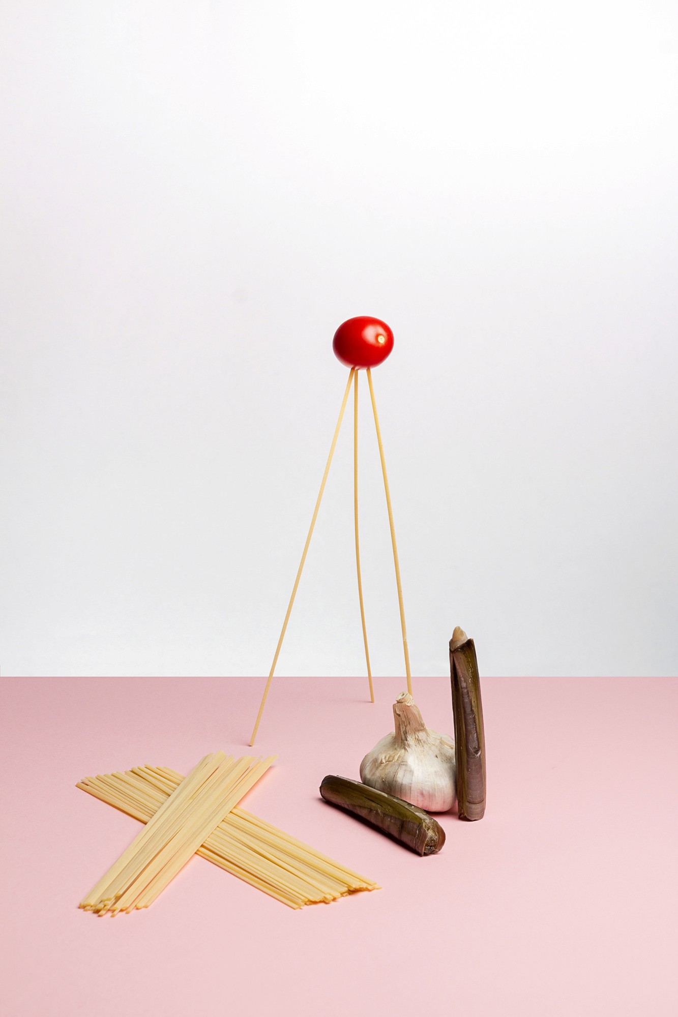 Nature morte studio sur une table avec un fond rose d'ingrédients mise en scène comme des spaghettis, une tomate cerise, des couteaux et une gousse d'ail