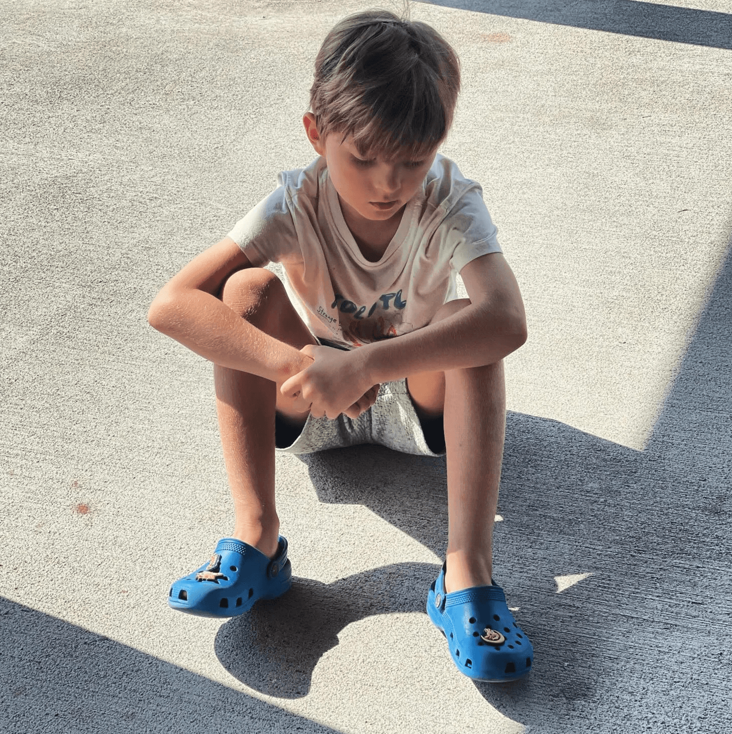 A young boy with disheveled hair and a frown sits on the ground, his shoulders slumped forward and a look of disappointment evident on his face. The boy's eyes appear downcast, reflecting a sense of sadness or frustration. The lighting in the image is dim, emphasizing the somber mood.