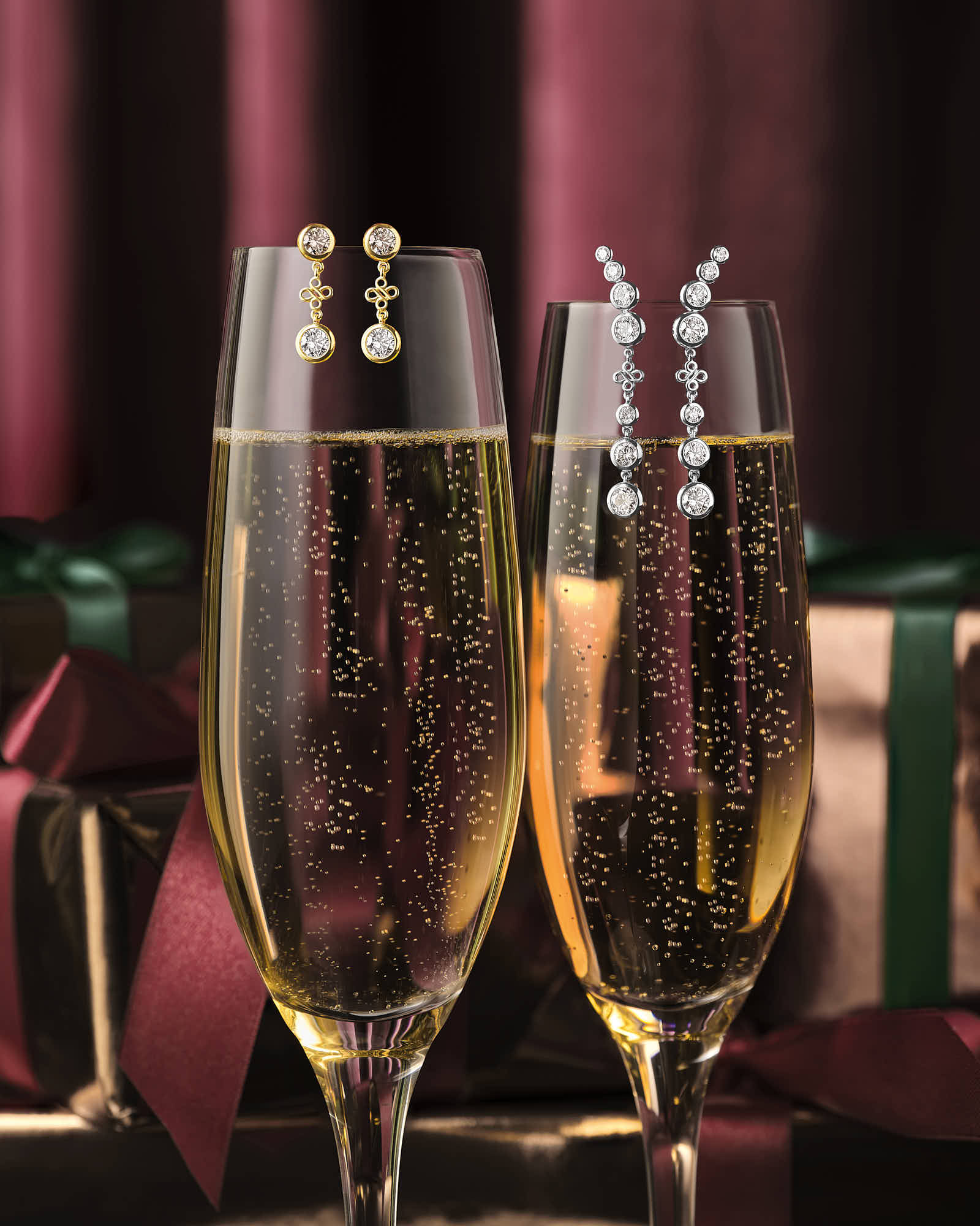 Two elegant champagne flutes with diamond earrings resting on them sit against a rich red backdrop, with gift boxes adorned in green and gold nearby.