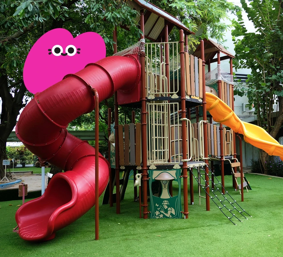 Colorful playground with tall structures, featuring a red spiral tube slide and an orange straight slide, set against lush green trees. Playful mood.