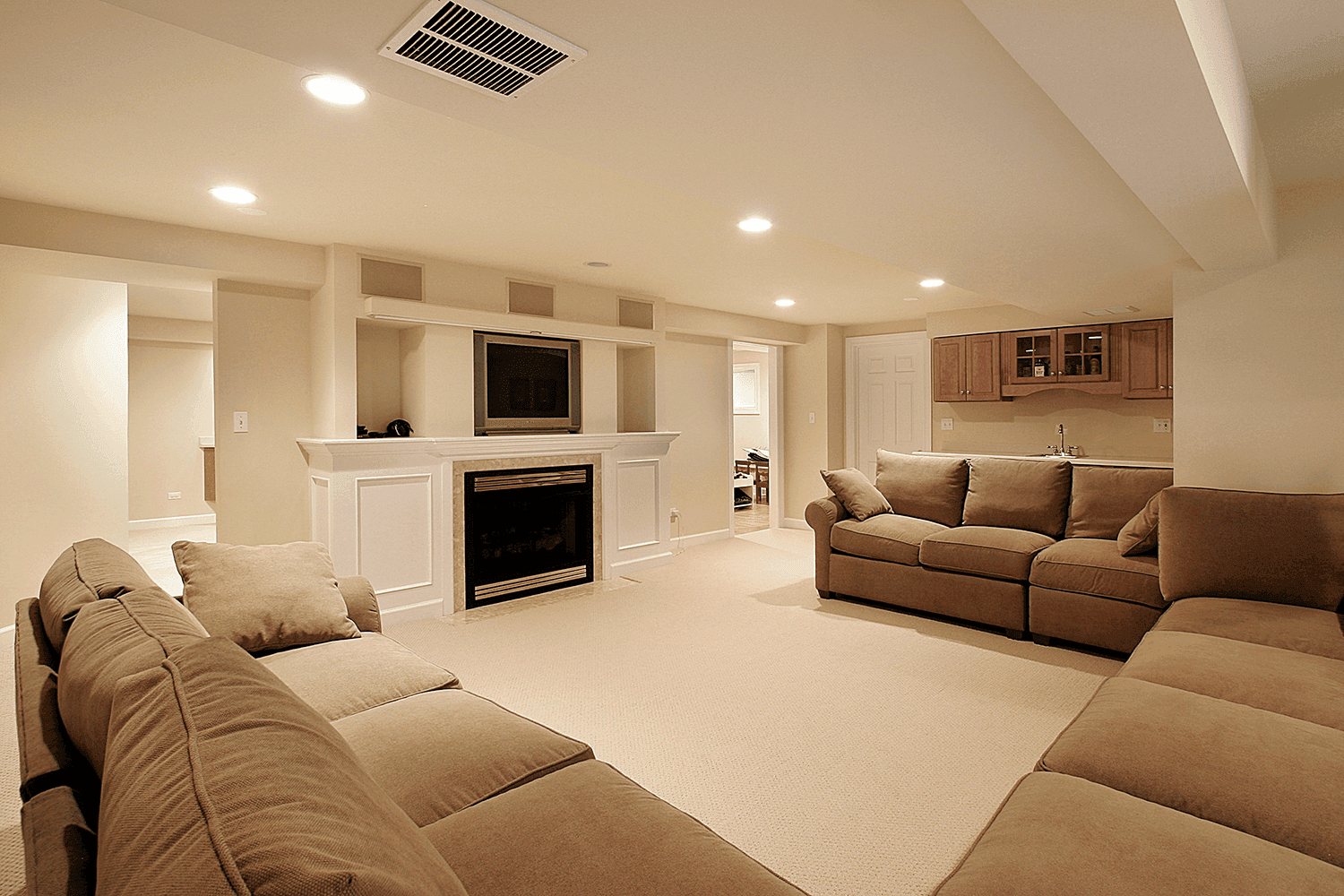 A traditional-style basement with beige carpet, large sectional sofas, and a built-in fireplace beneath a mounted TV. The room includes a small kitchenette with wooden cabinets and recessed lighting for a warm, inviting atmosphere.