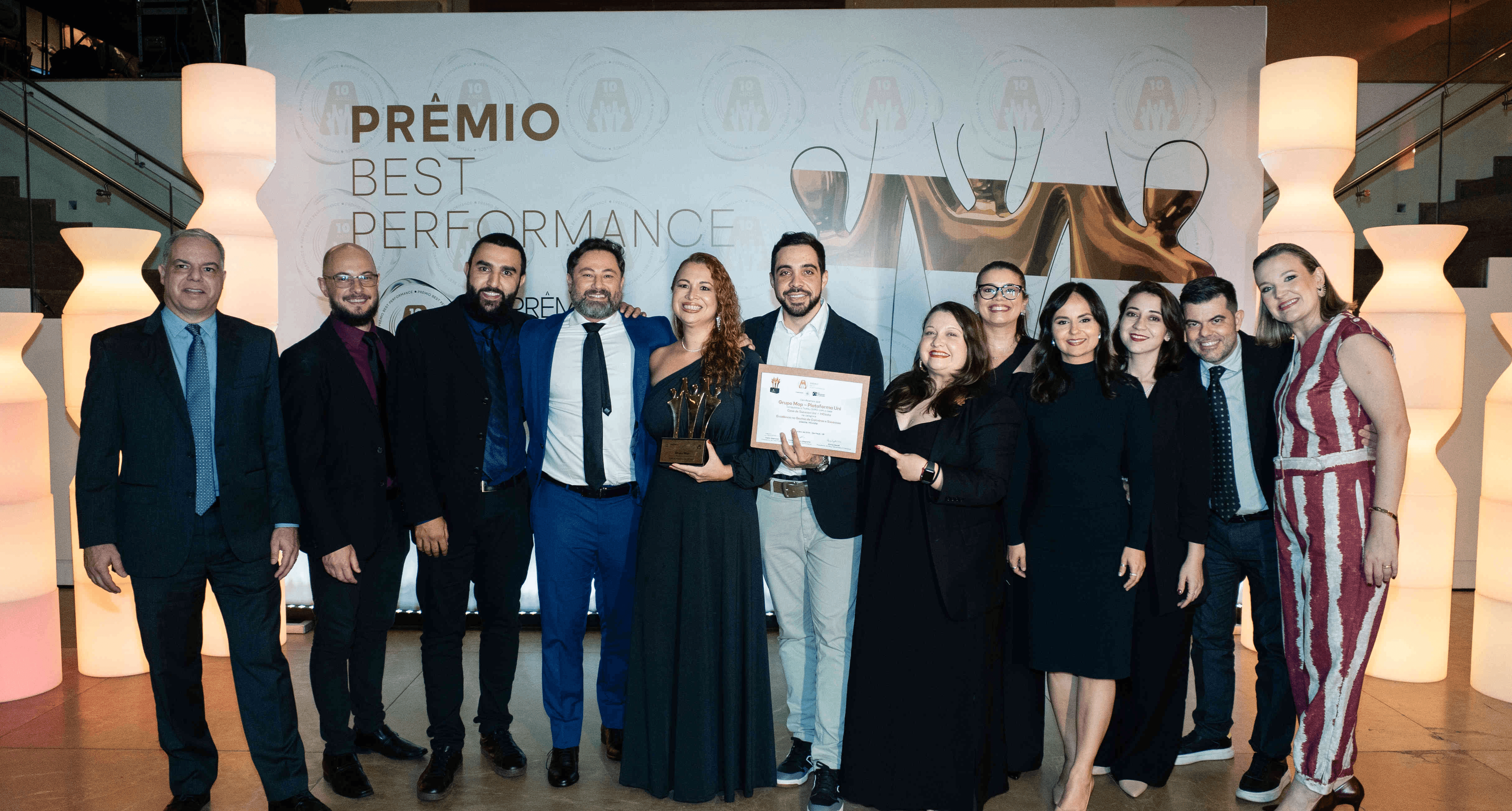 Executivos do Grupo Mop posando com troféu ouro e certificado no palco do Prêmio Best Performance 2025