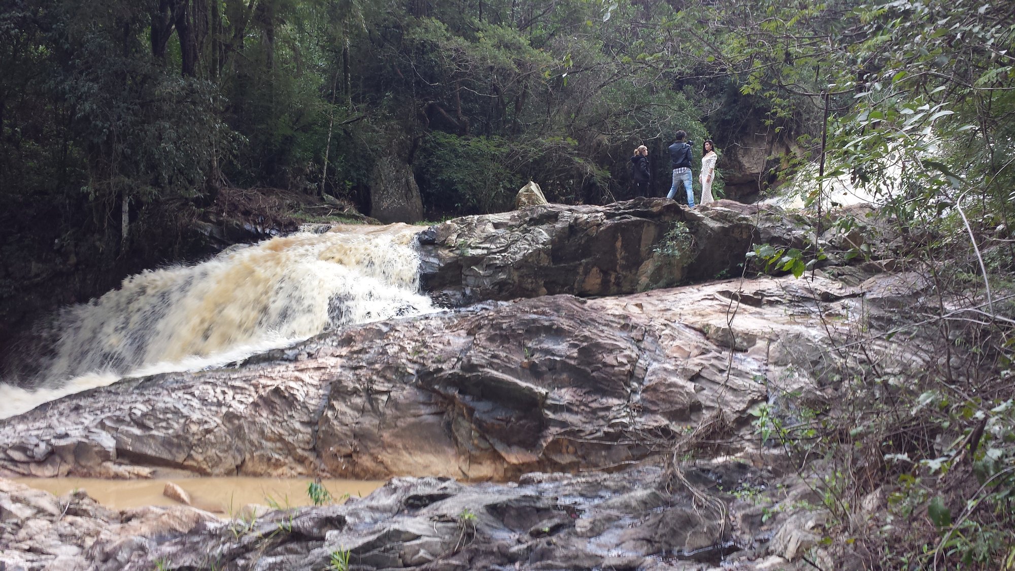 Cachoeira sobre rochas na mata com pessoas no alto das pedras, making of com a atriz Glenda Poletto