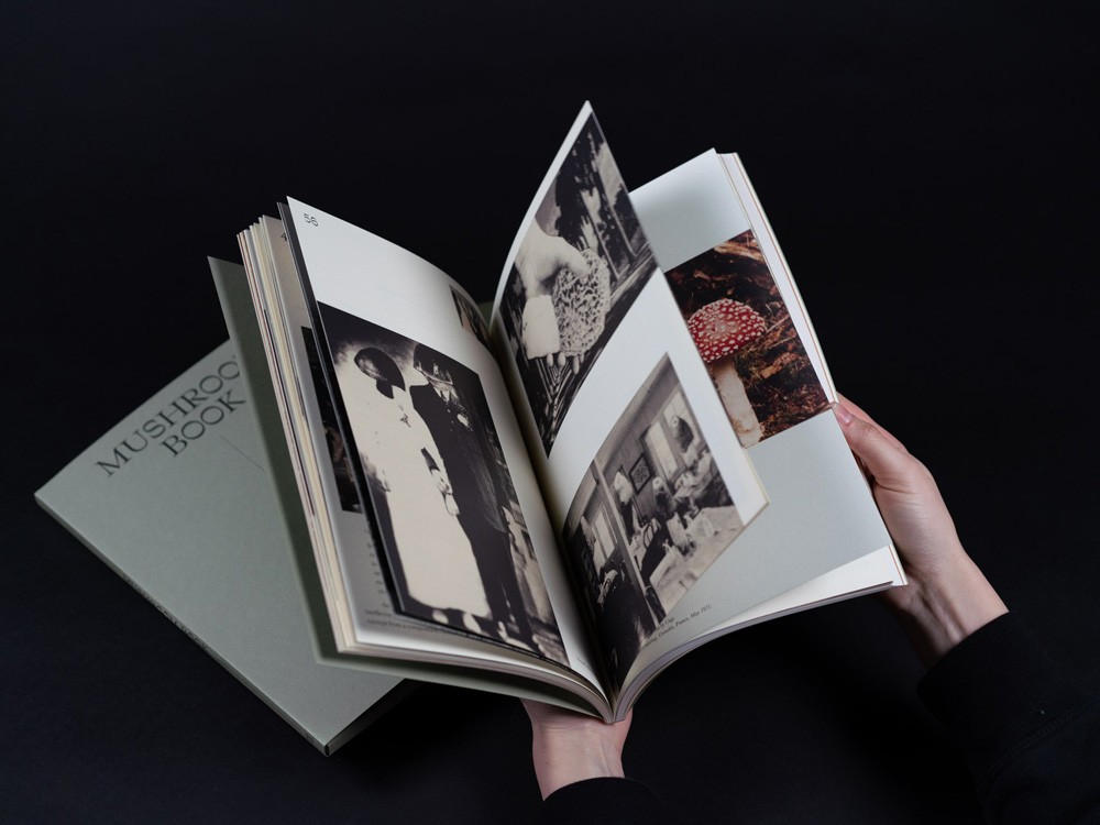 Een close-up foto van een opengeslagen boek dat met beide handen wordt vastgehouden tegen een zwarte achtergrond. De linkerpagina toont een zwart-wit portret van een stel, terwijl de rechterpagina's doorbladeren en een kleurrijke afbeelding van een vliegenzwam onthullen.