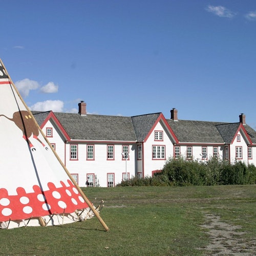 A decorated tent stands on a grassy field with a large, multi-story white building with red trim in the background under a clear blue sky.