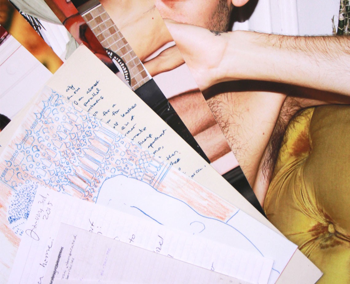 The image shows a close-up of a person's arm resting on a surface covered with various papers and drawings. The arm is positioned near a drawing that appears to be a blueprint or sketch of a structure, possibly a house or a room, with lines and shapes indicating walls and floor plans. Other papers are visible underneath and around the drawing, some with handwritten text and markings. The person has a tattoo on their arm and is wearing a mustard-yellow or gold-colored top. The background is out of focus and does not provide clear details about the setting.