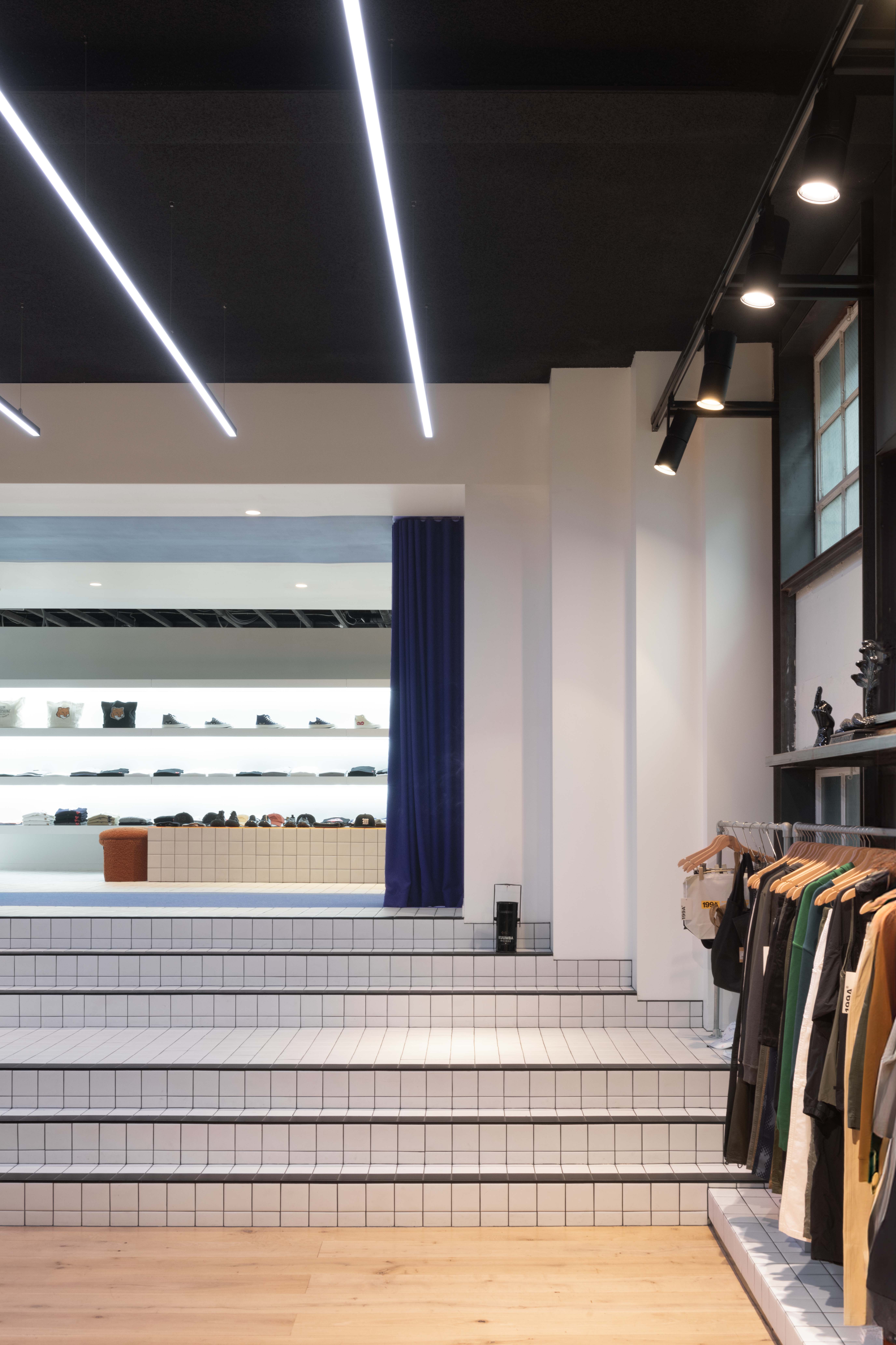 Vertical interior view of the 199A® store highlighting the tiled staircase stage, linear ceiling lights, deep blue curtain backdrop, and a side wall of hanging garments under track lighting.