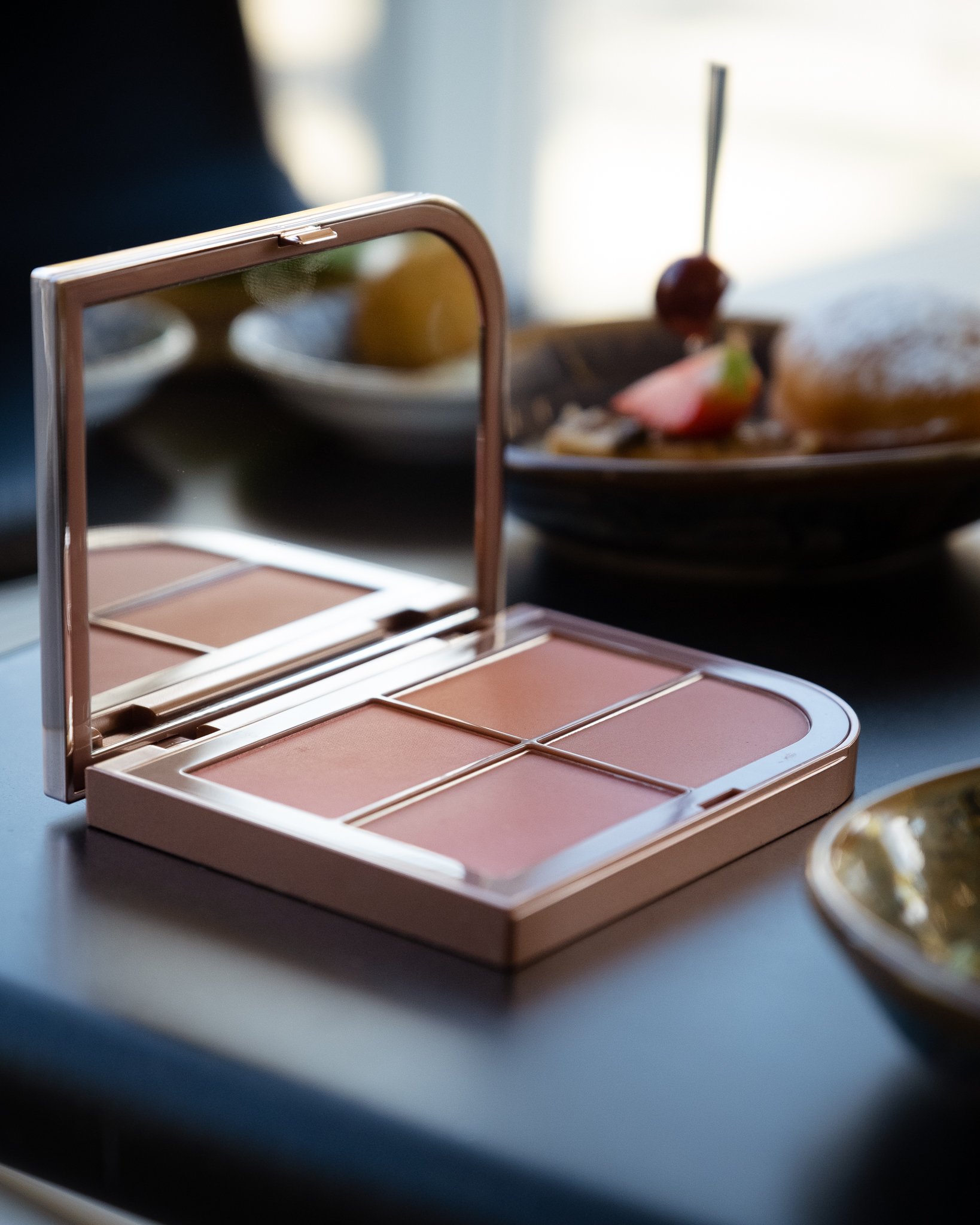 Open makeup palette with four shades of pink on a dark surface, with plates of food in the background.