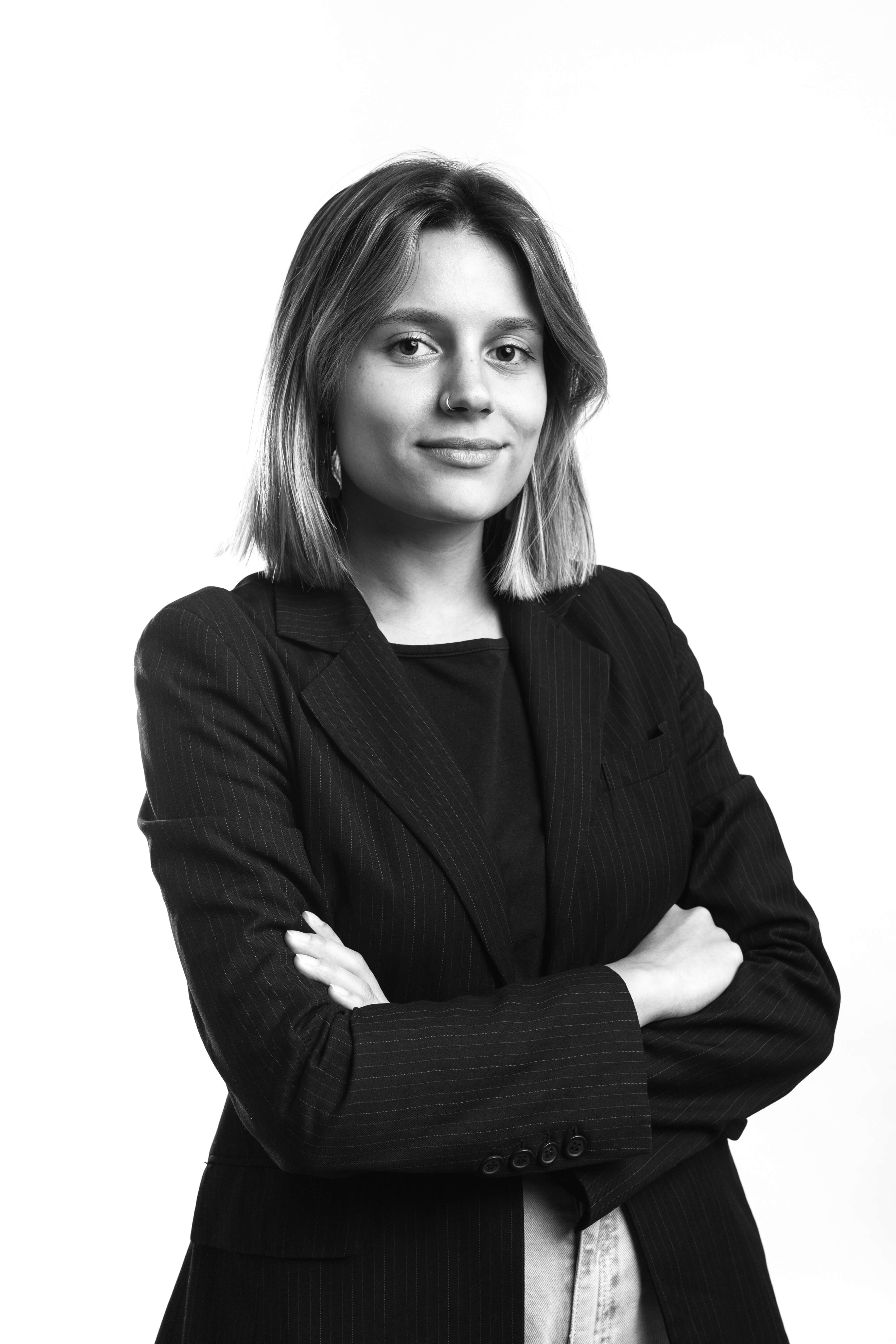 Black and white portrait of a person with medium-length hair, wearing a pinstripe blazer, arms crossed, smiling confidently against a white background.