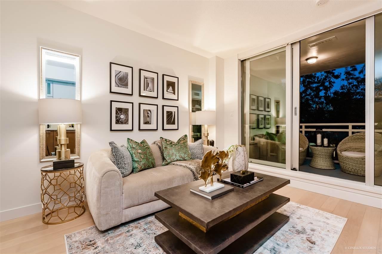 A modern living room with a cozy sofa, decorative cushions, a coffee table, and large windows showing greenery outside.