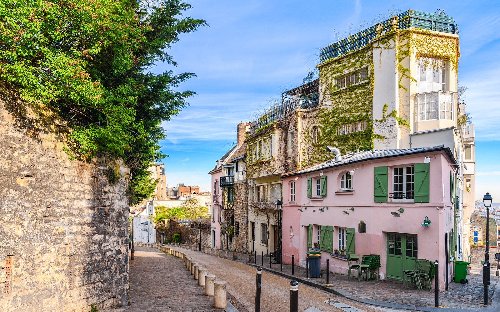 Brosteinbelagt gate i Montmartre, Paris, omgitt av bygninger dekket med eføy og en rosa kafé.