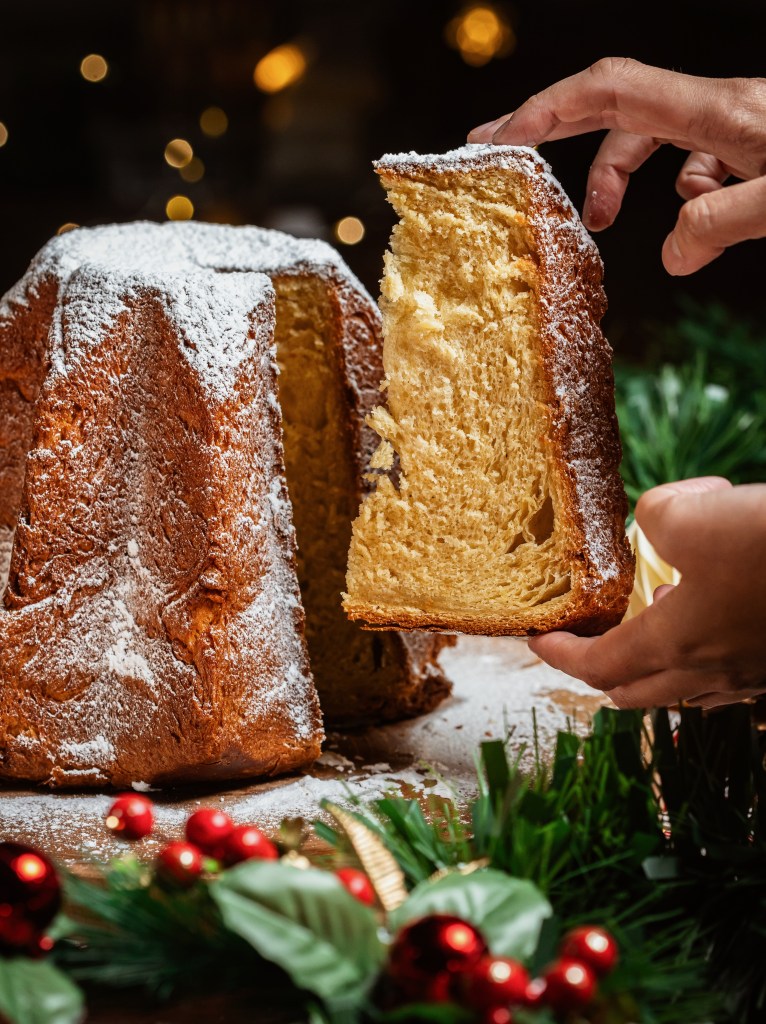 A hand reaching for a slice of a festive cake dusted with powdered sugar, surrounded by Christmas decorations like red ornaments and greenery.
