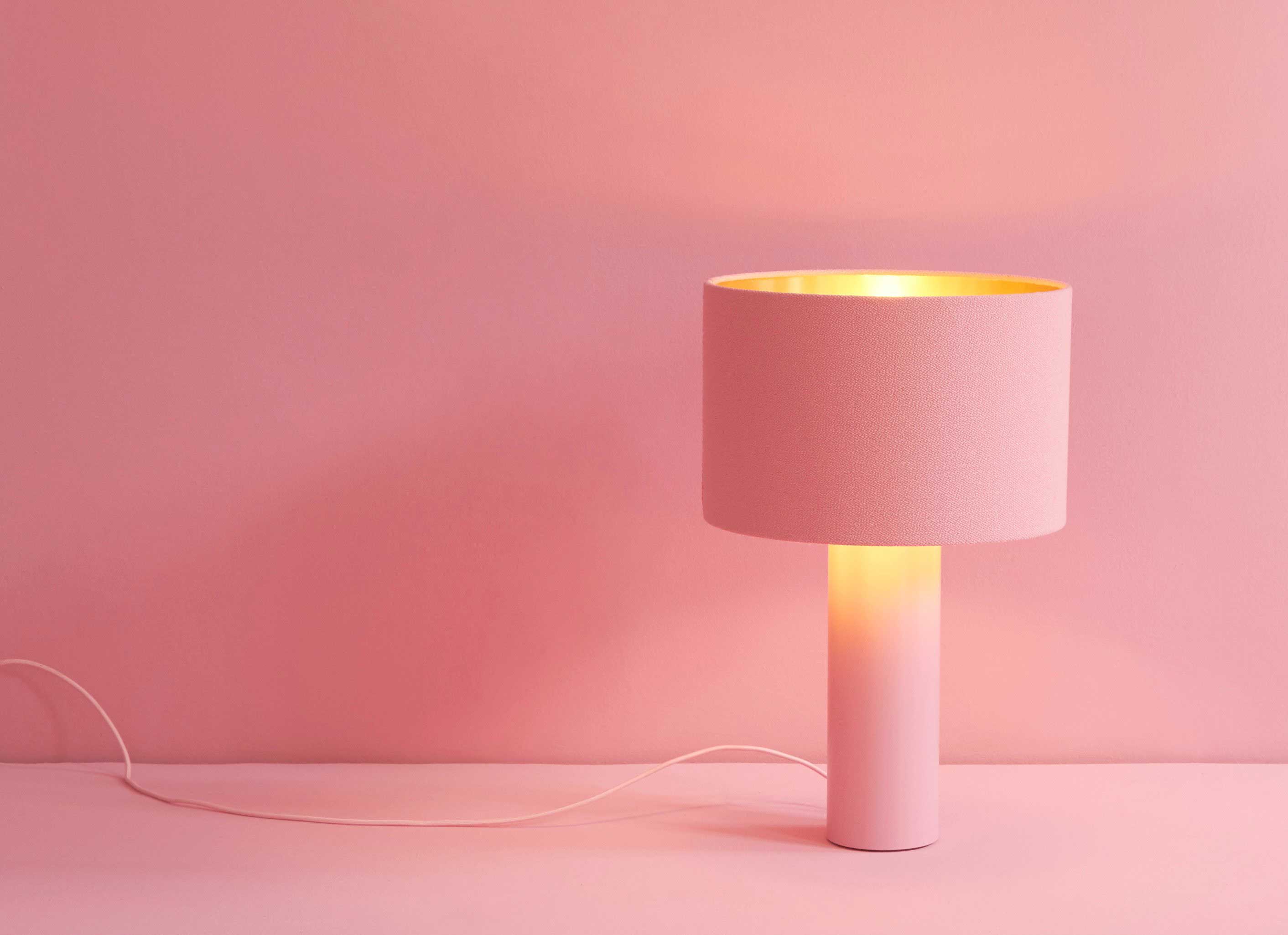 A single modern table lamp with a light pink cylindrical base and a cylindrical lampshade. The light glowing from the lamp creates a warm, pinkish hue against a matching pink background.