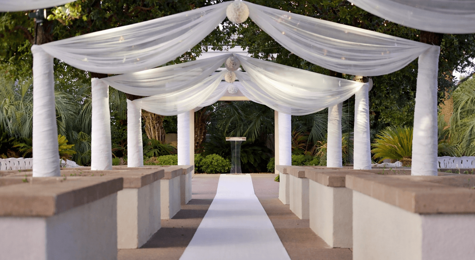 Outdoor restaurant terrace overlooking a tranquil lake at sunset. White tables and chairs are set with glassware. Sheer curtains add elegance. Calm ambiance.