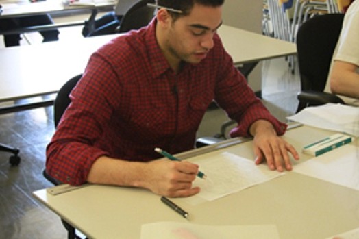 A photo of a man sitting at a desk and drawing a sketch on a piece of paper.