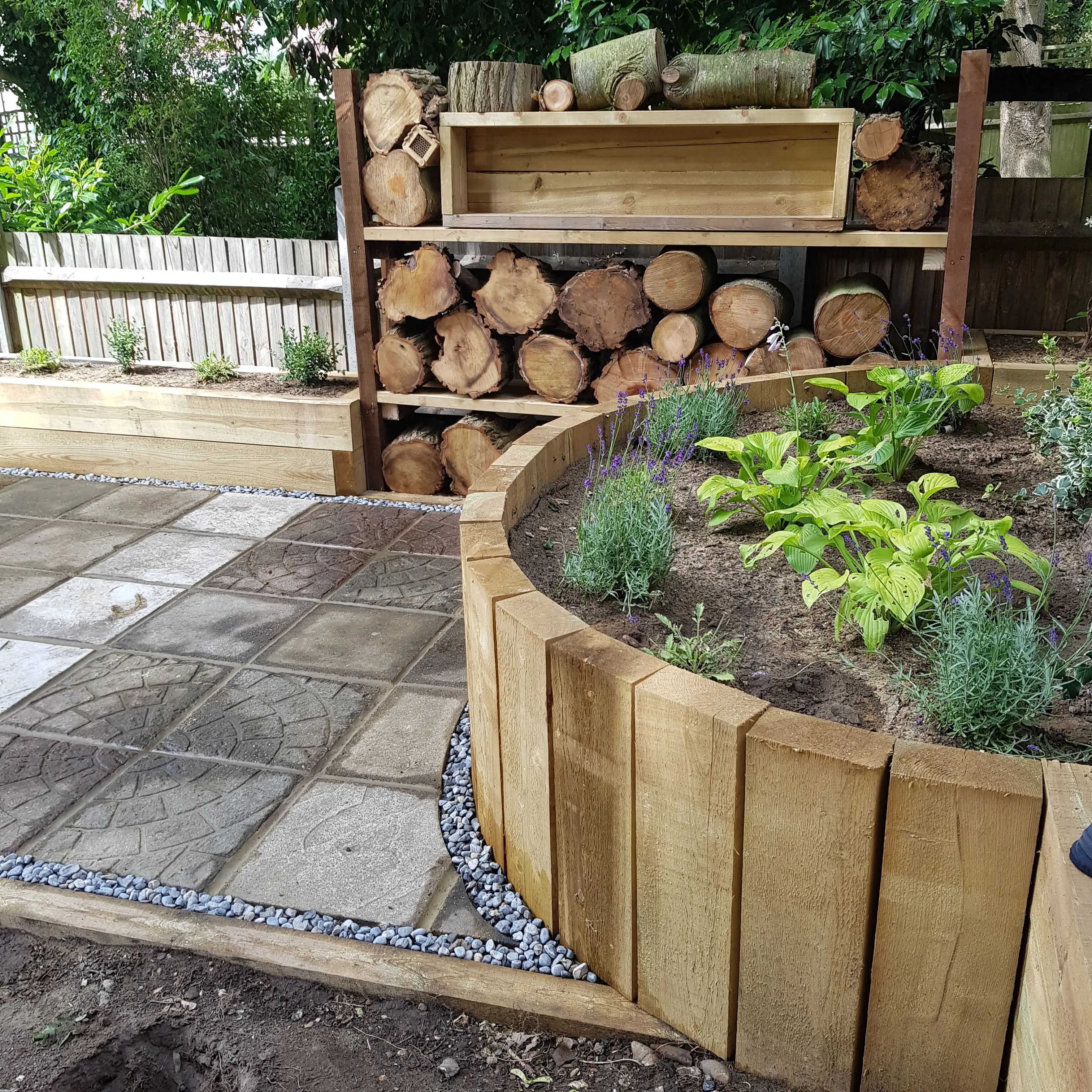 A landscaped garden featuring wooden planters, stone pathways, and stacked logs in a serene outdoor setting.
