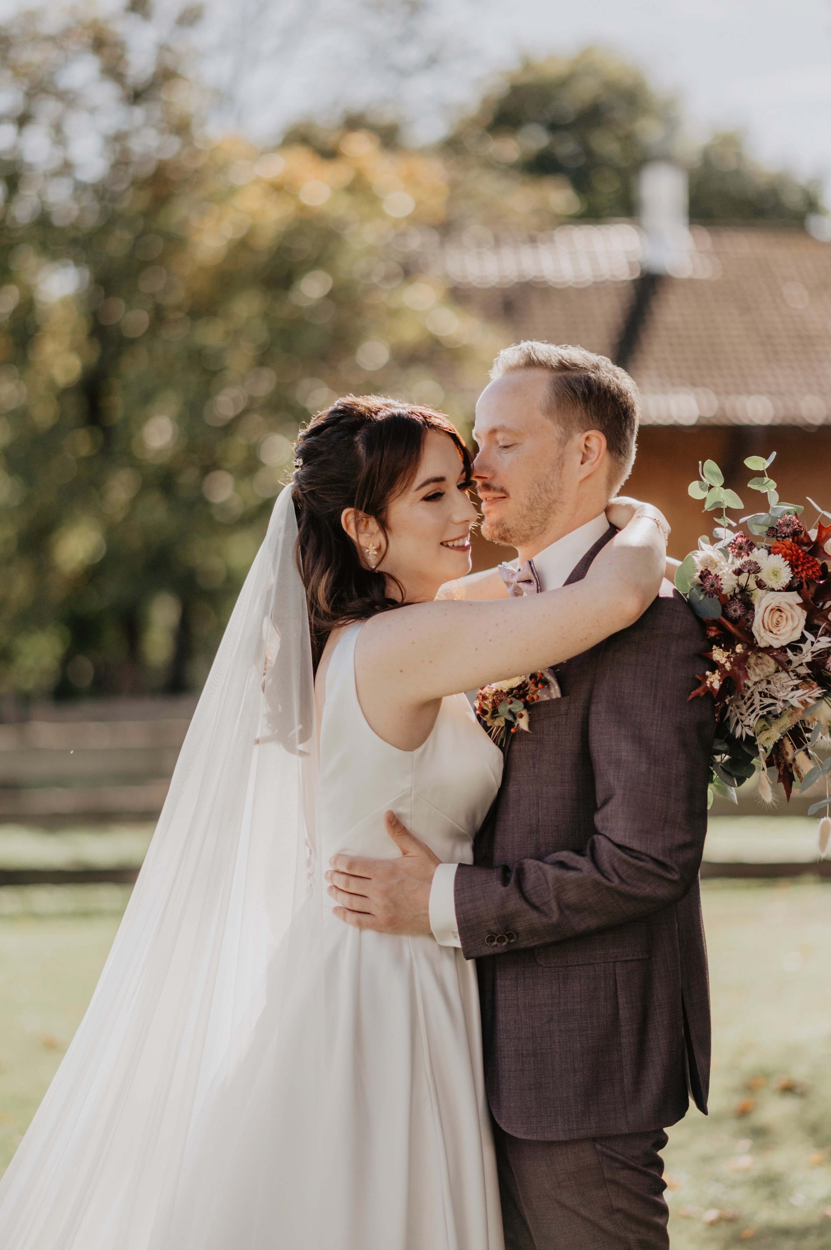 Ein frisch verheiratetes Paar umarmt sich liebevoll im Freien. Die Braut trägt ein elegantes, weißes Kleid mit Schleier, und der Bräutigam einen stilvollen, dunkelgrauen Anzug mit floraler Ansteckblume. Sie blicken sich glücklich an, während die Braut einen herbstlichen Brautstrauß mit warmen Farbtönen hält. Im Hintergrund verschwimmen Bäume und ein Gebäude in weichem Sonnenlicht.Kundenfeedback Hochzeitspaar für Maria Wiethaler freie Rednerin und Sängerin