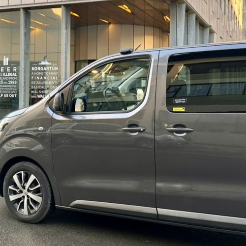 Una minivan gris estacionada en la calle frente a un edificio con fachada de vidrio con letreros y texto visibles a través de las ventanas.