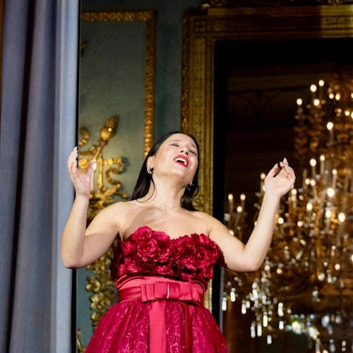 一名身穿綴有玫瑰裝飾的紅色抹胸連身裙的女子，在華麗的房間內深情高歌，背景有一盞大型吊燈。