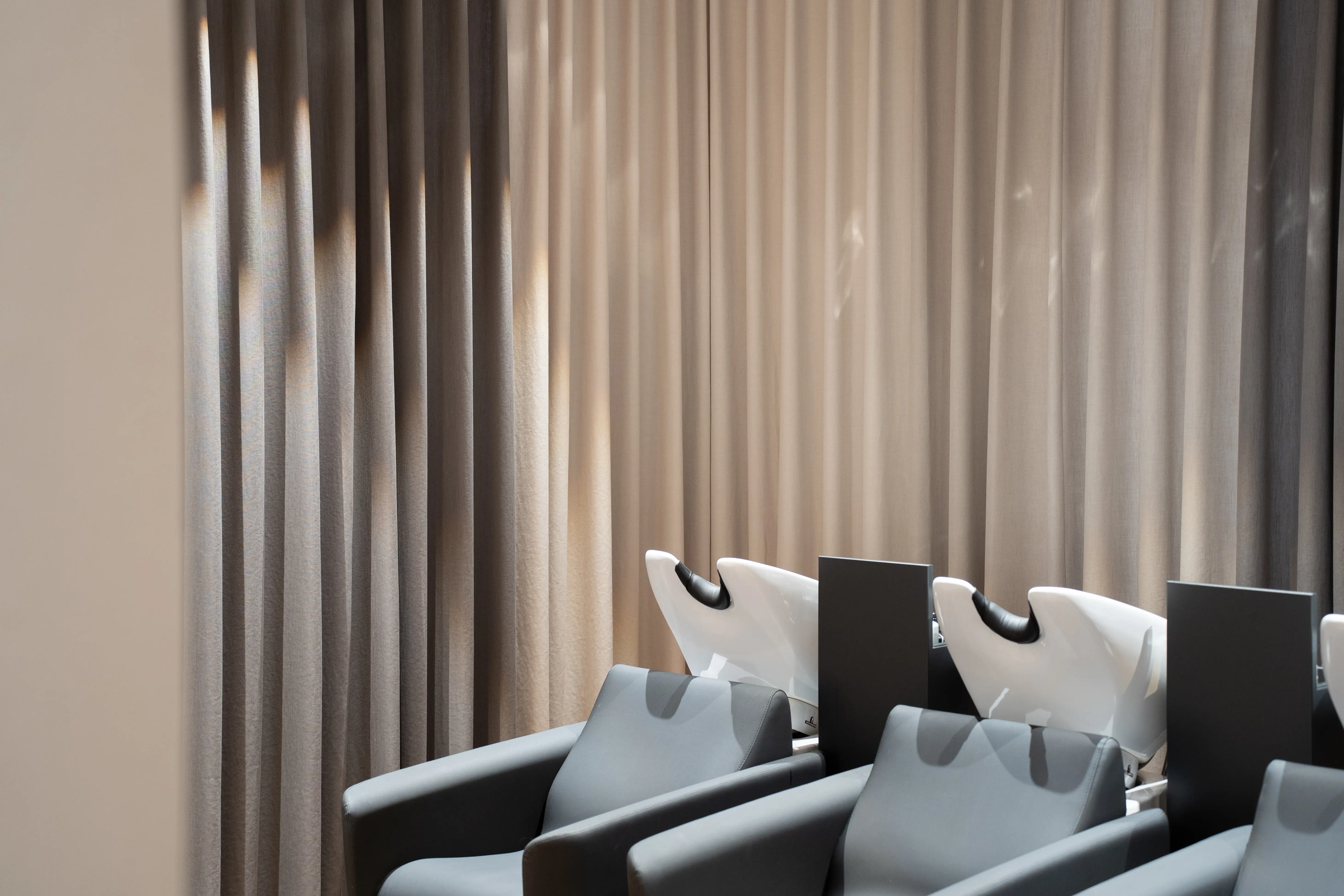 Detail of white ceramic backwash basins on grey-blue lounge chairs at EMPL FRISEURE in front of softly lit beige curtains.