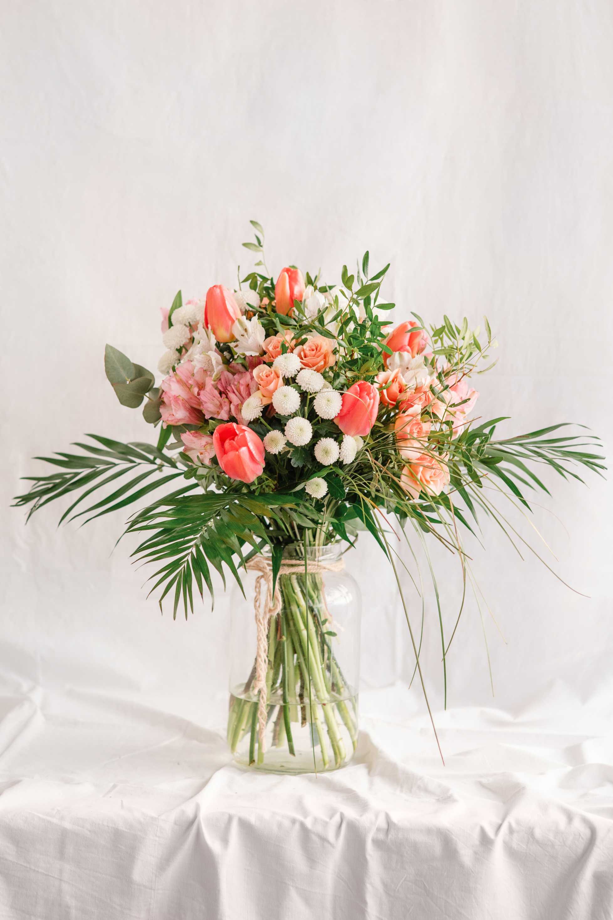 Ram de flors fresques en tons rosats, corals i blancs amb tulipes, roses, crisantems i verdures ornamentals, presentat en un gerro de vidre amb corda de jute.