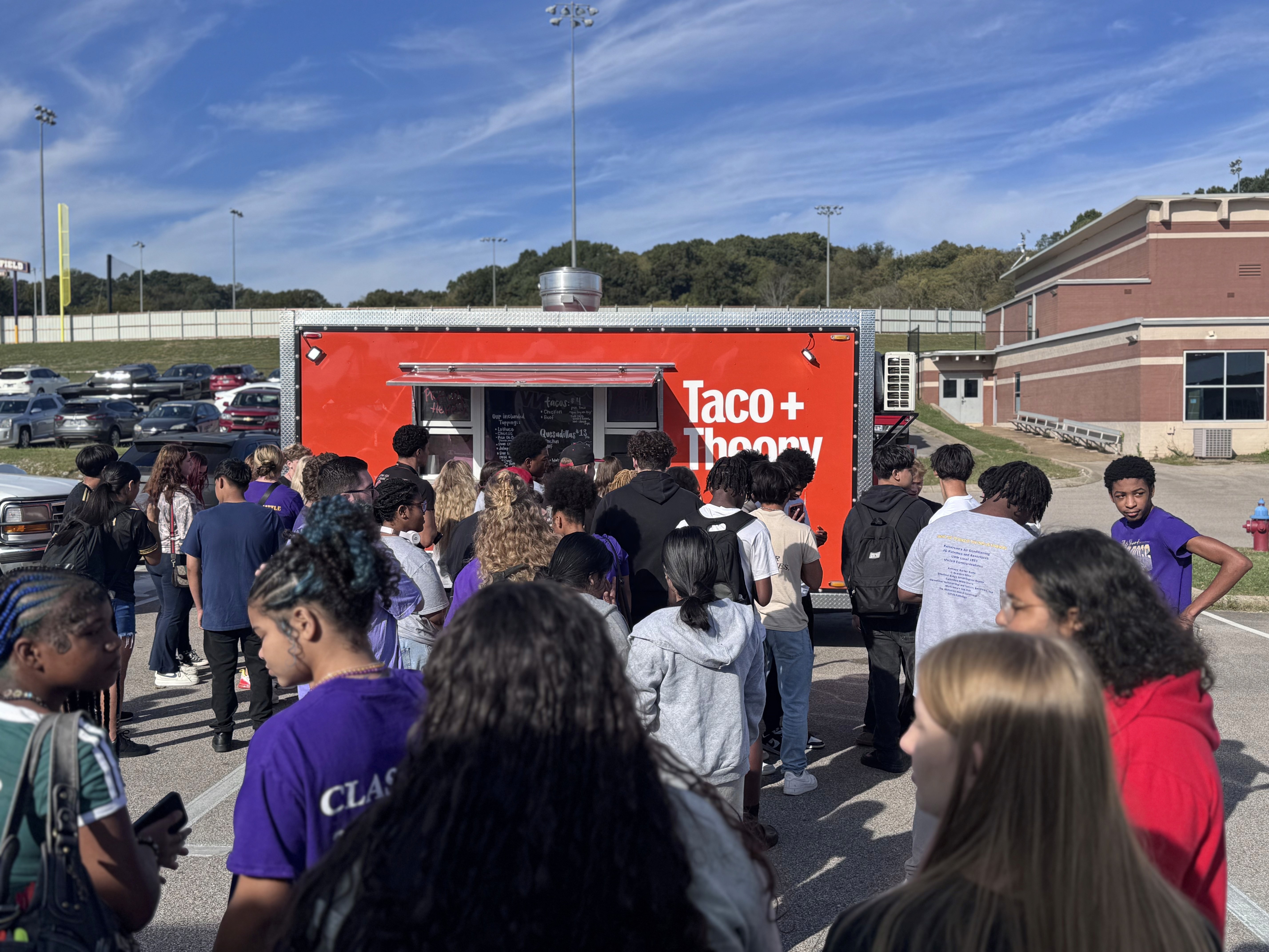 A bright, modern dining room featuring a long light wood table set with several matching wooden chairs and white cushions, illuminated by natural light streaming through large windows overlooking a lush green and sunlit yard.