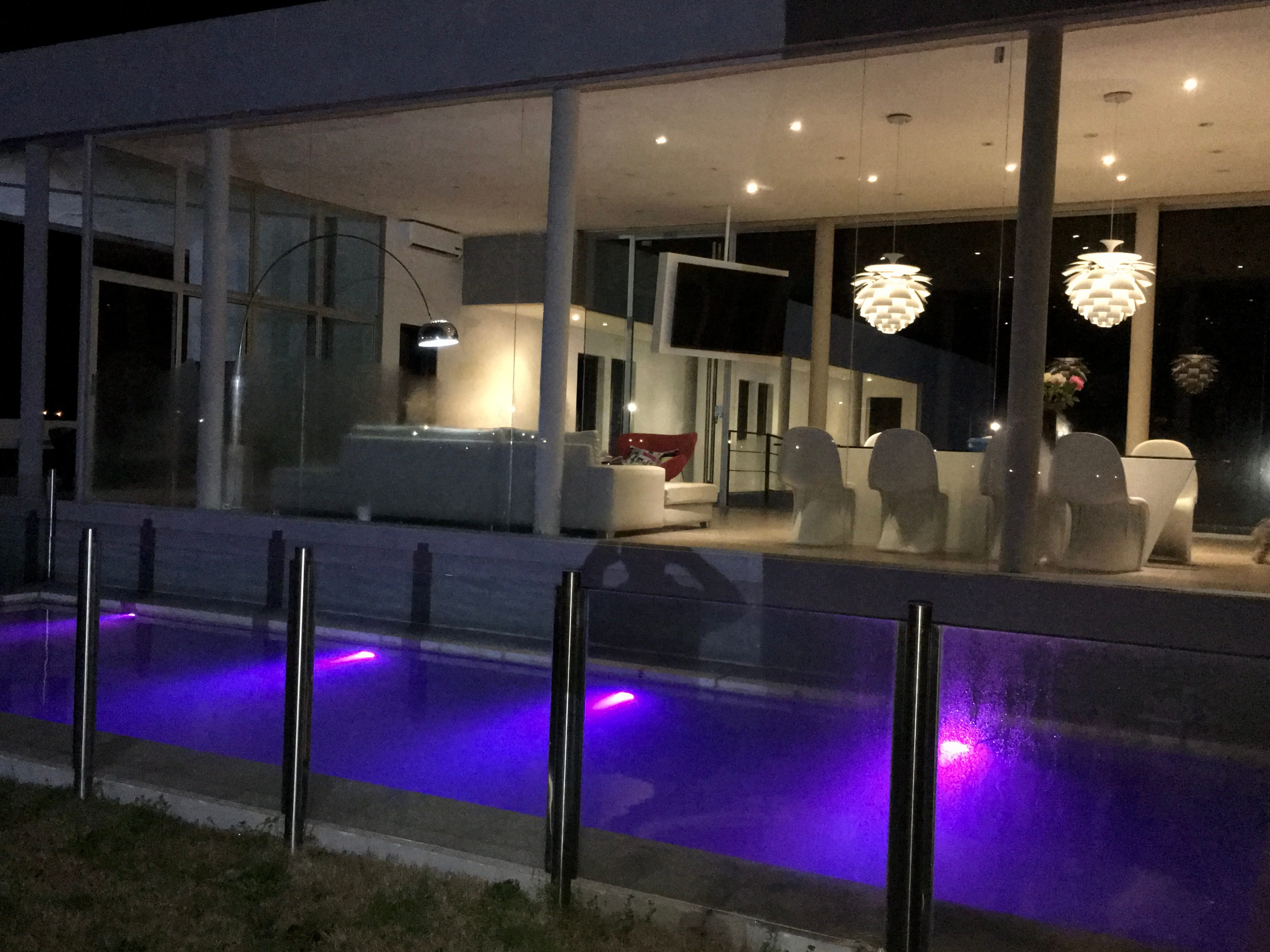 Espectacular piscina iluminada con LED violeta de noche, ubicada frente a una casa de arquitectura moderna con grandes ventanales iluminados y cerco de vidrio.
