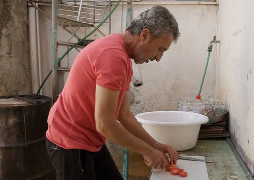 Άνδρας με κόκκινη μπλούζα κόβει ντομάτες σε μικρή κουζίνα με άσπρους τοίχους και μεταλλικά αντικείμενα.