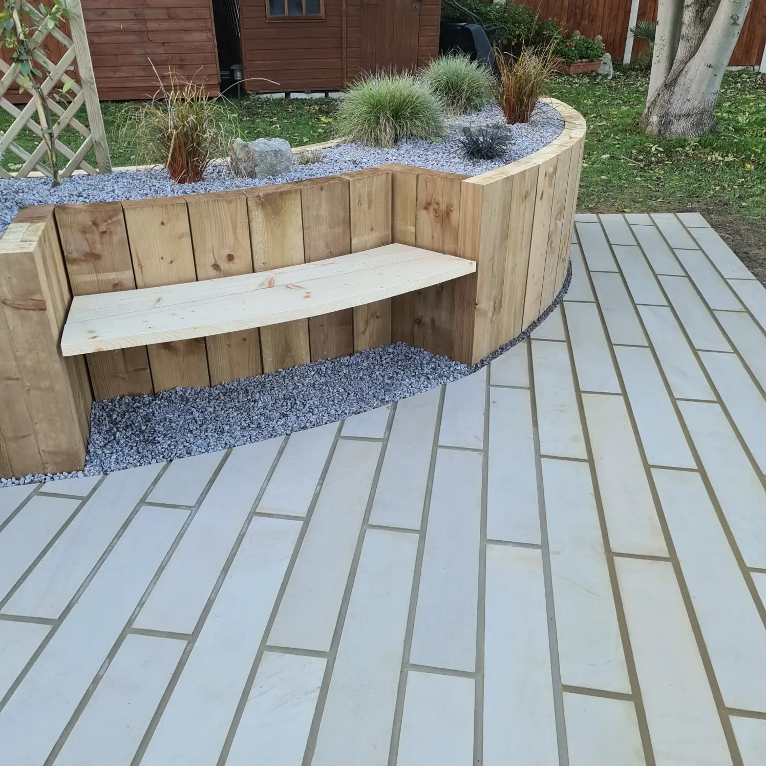 A curved wooden bench surrounded by plants on a paved outdoor patio area.