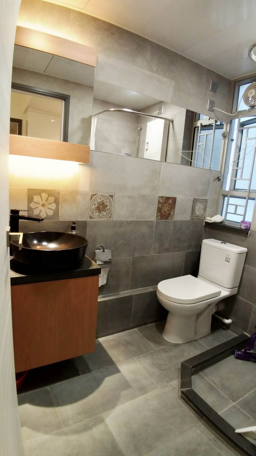 This modern bathroom features a sleek black vessel sink paired with decorative accent tiles.Vanity Details: The bathroom includes a black vessel sink installed on a dark countertop with a wooden cabinet underneath.Wall Design: The walls are tiled with large grey ceramic tiles, accented by a row of lighter decorative tiles featuring floral and patterned motifs.Fixtures: A modern, matte black faucet is mounted on the wall or vanity, and a standard white toilet sits next to the sink