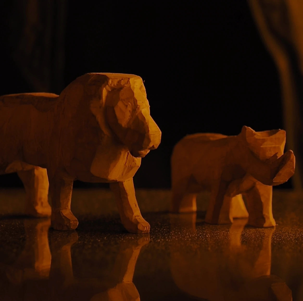 Two roughly carved wooden animal figurines, a lion and a smaller animal, reflected on a dark surface against a black background, in amber and orange