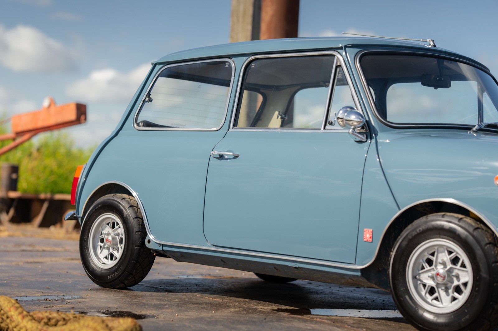 A light blue vintage car parked outdoors with a colorful vehicle and cloudy sky in the background.